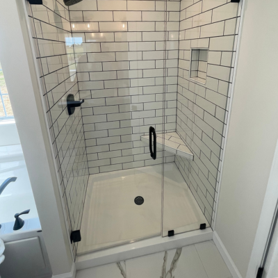 Glass-enclosed walk-in shower with white subway tile walls, a black showerhead and controls on the left, and a clear glass door over a tiled shower base with a small floor drain.